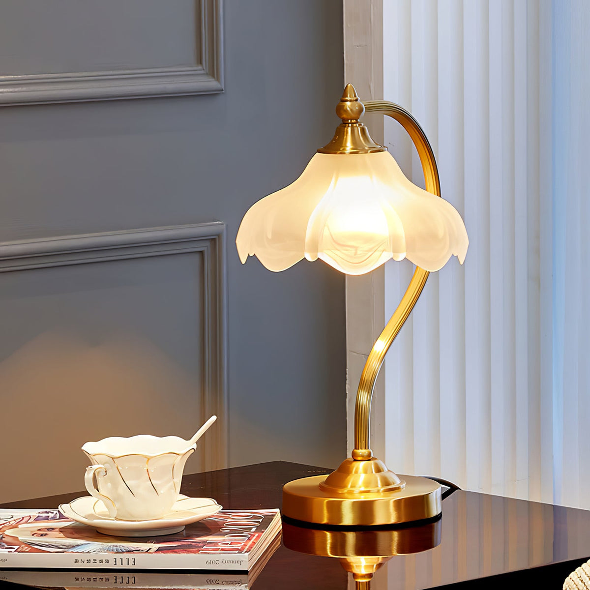 A lit Clérachic table lamp on a glossy dark table, with a white cup and saucer and a stack of magazines, against a blue-grey panelled wall.