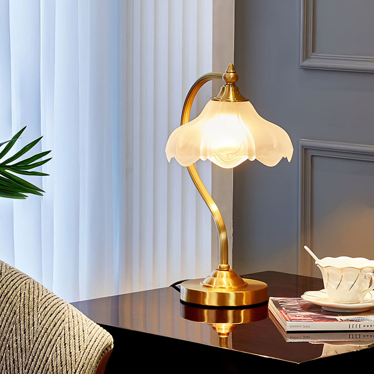 A Clérachic lamp on a polished dark table by a window with sheer curtains, a teacup, magazines and a striped cushion beside it.