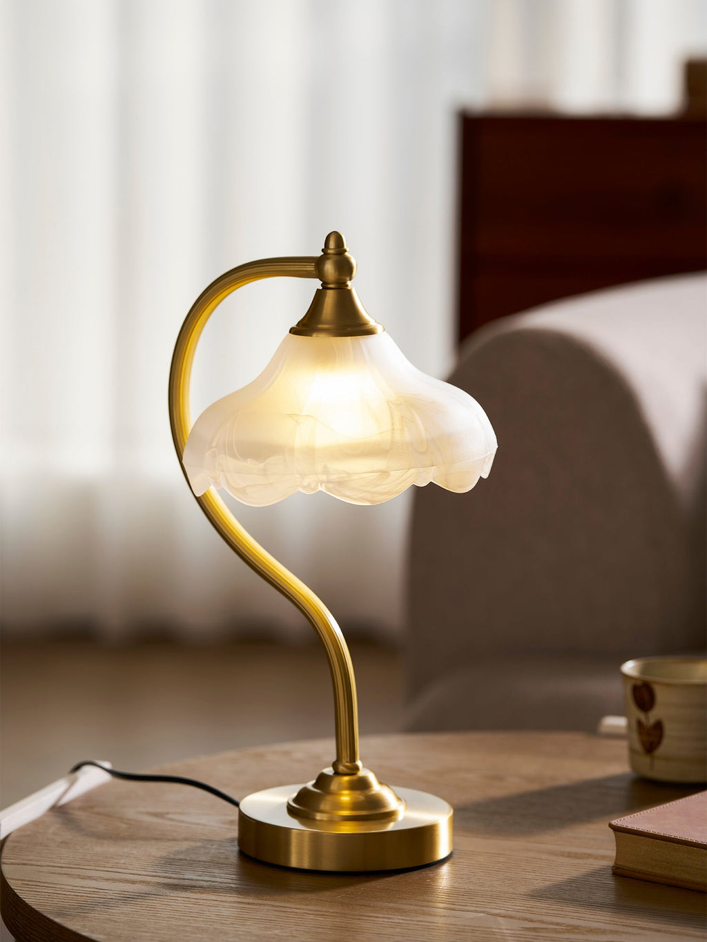 Low view of this Clérachic lamp on a round wooden table, with a mug and a book to the right and a soft grey sofa behind.