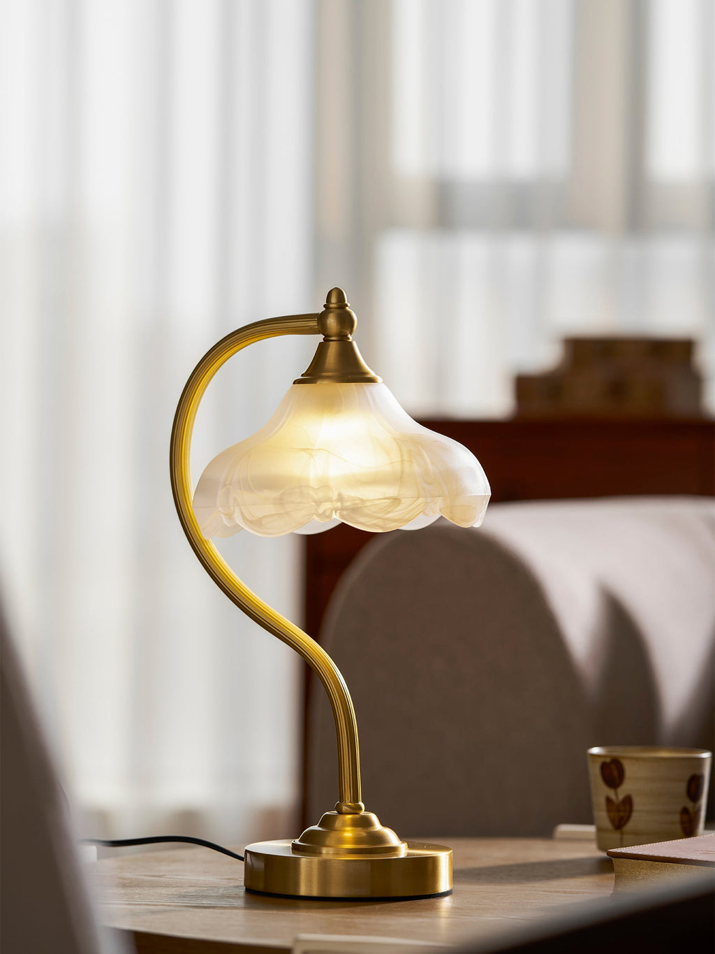 This Clérachic lamp on a wooden table with a small patterned mug, with sheer curtains and a grey sofa softly blurred behind.