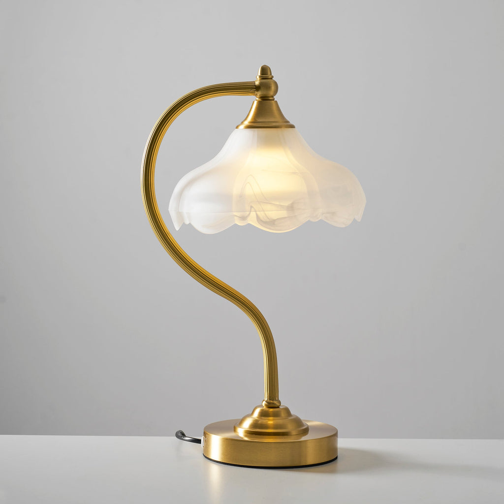 A Clérachic lamp on a white surface in front of a grey wall, with the bulb glowing inside the frosted scalloped shade.