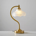 A Clérachic lamp on a white surface in front of a grey wall, with the bulb glowing inside the frosted scalloped shade.