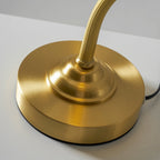 Close view of the round brushed gold base, the stepped collar and the ribbed arm of this Clérachic lamp, with the black cord at the edge.