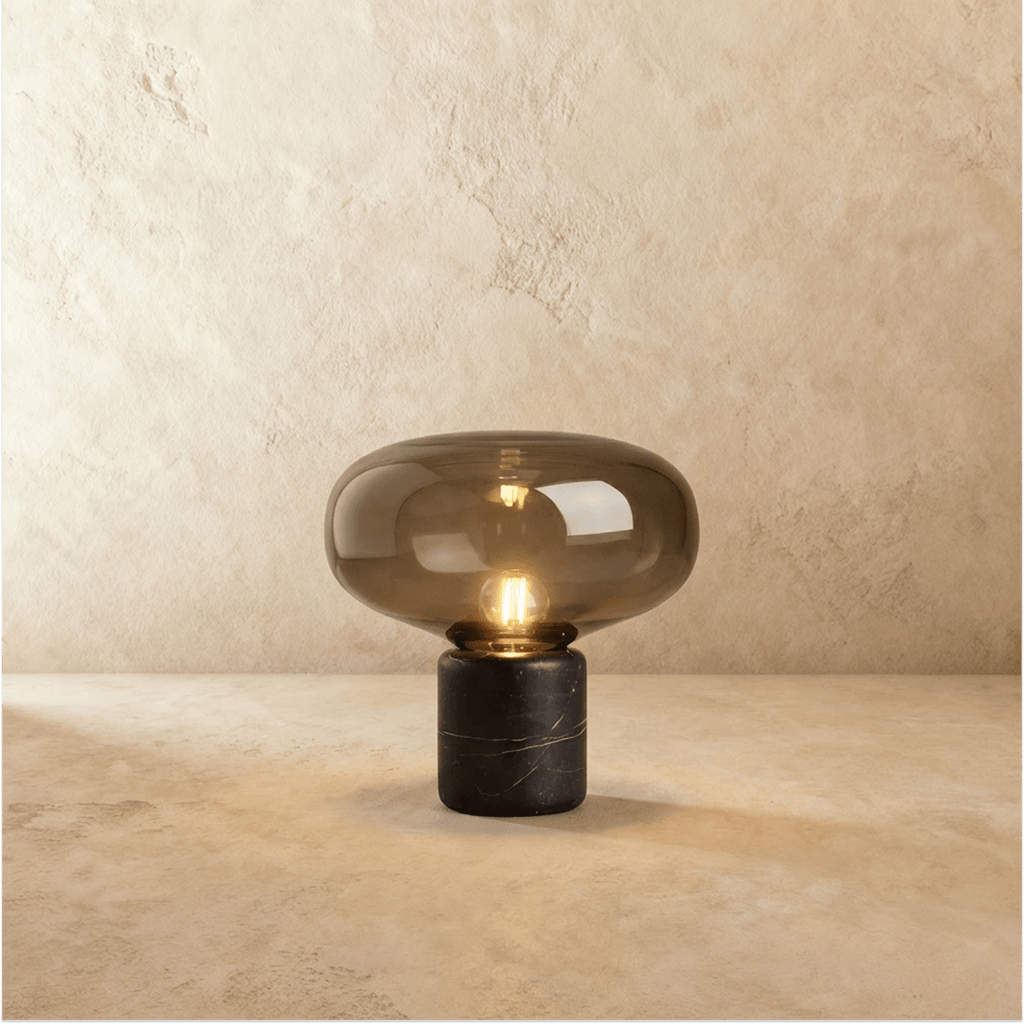 Clérachic table lamp with a wide smoked glass shade over a black marble base, filament bulb lit, on warm plaster