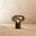 Clérachic table lamp with a wide smoked glass shade over a black marble base, filament bulb lit, on warm plaster