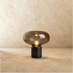 Clérachic table lamp with a wide smoked glass shade over a black marble base, filament bulb lit, on warm plaster