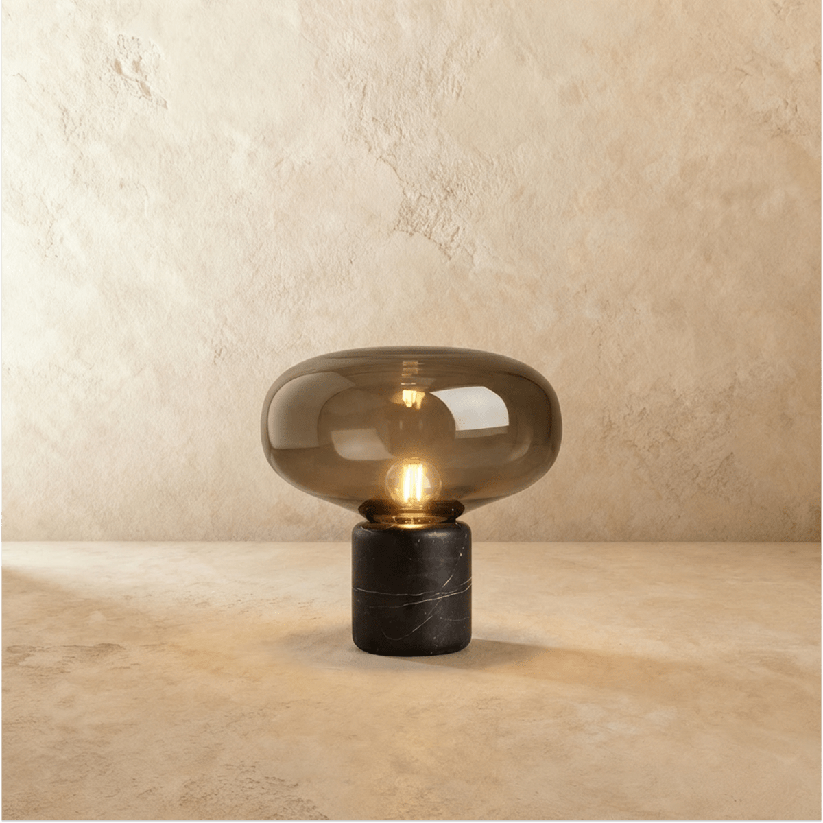 Clérachic table lamp with a wide smoked glass shade over a black marble base, filament bulb lit, on warm plaster
