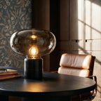 Clérachic table lamp lit warm on a black desk, its smoked glass shade and veined marble base by a patterned wallpaper