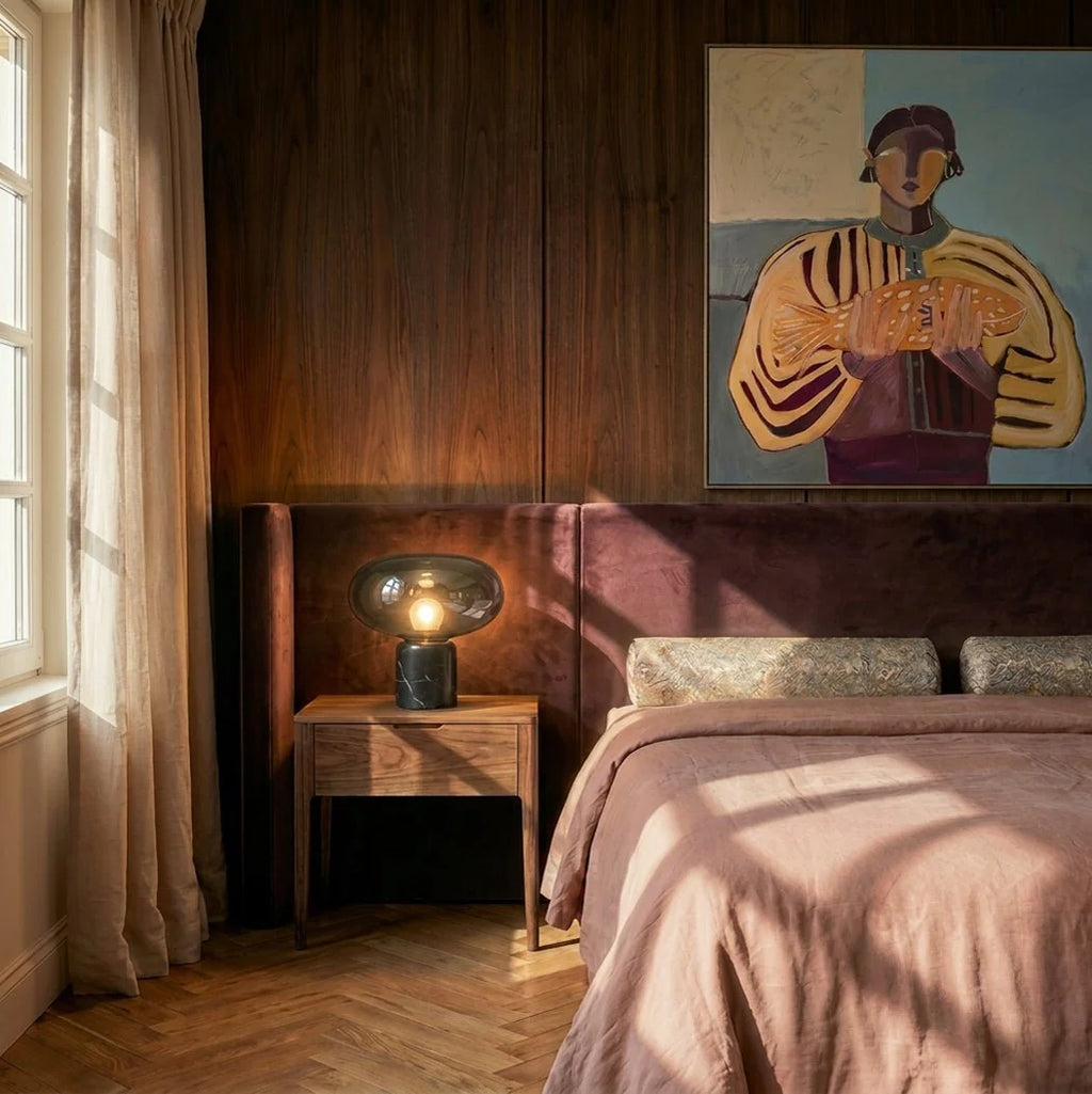Clérachic smoked glass table lamp glowing on a walnut bedside table beside a velvet headboard and framed painting