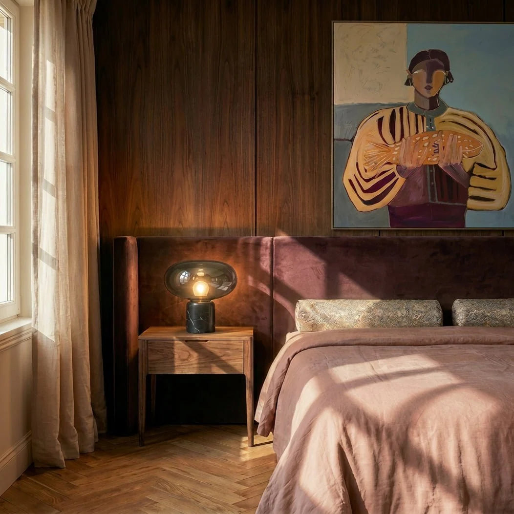 Clérachic smoked glass table lamp glowing on a walnut bedside table beside a velvet headboard and framed painting