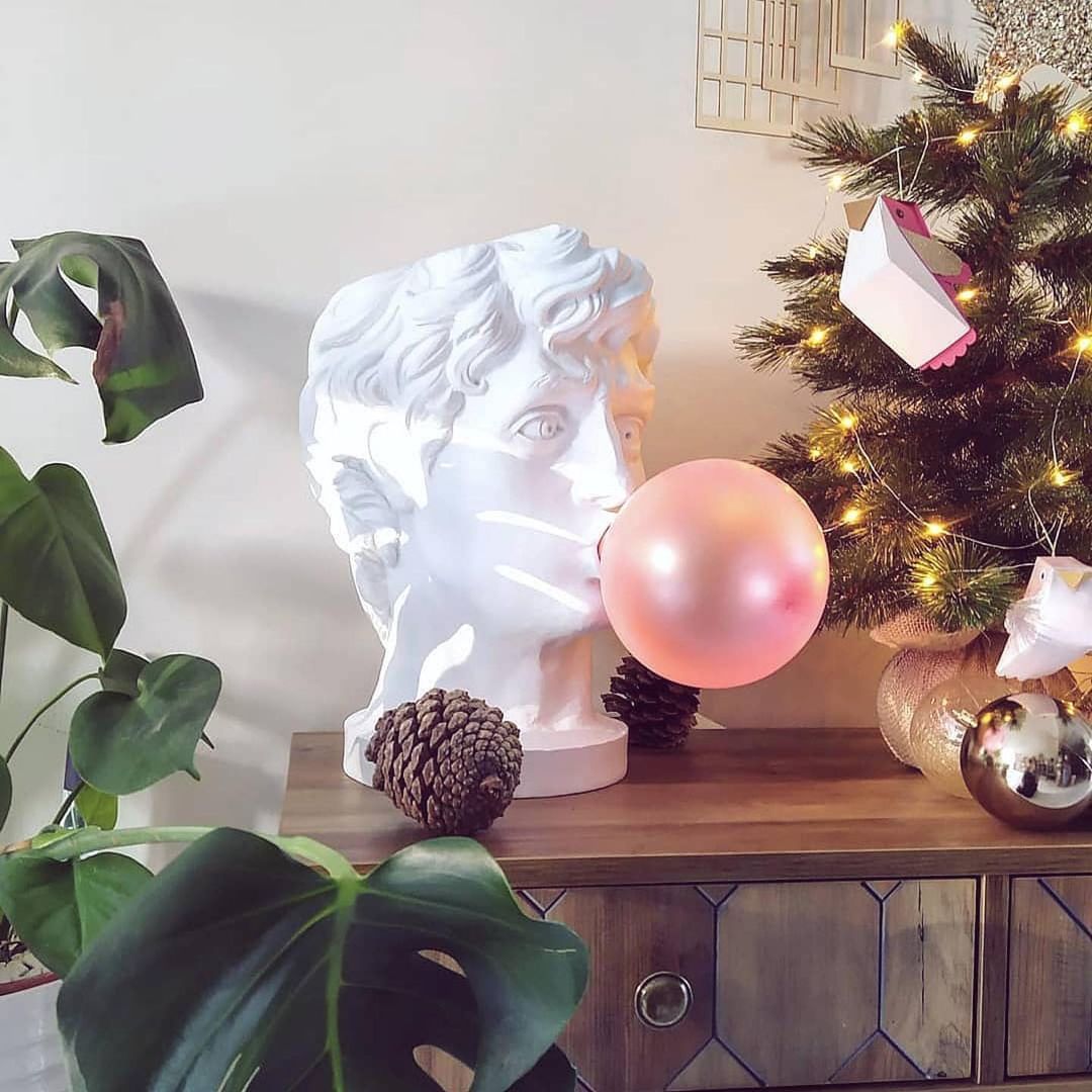 A white bust lamp from Clérachic blowing a pearly pink bubble, on a wooden console beside a monstera and a lit Christmas tree.