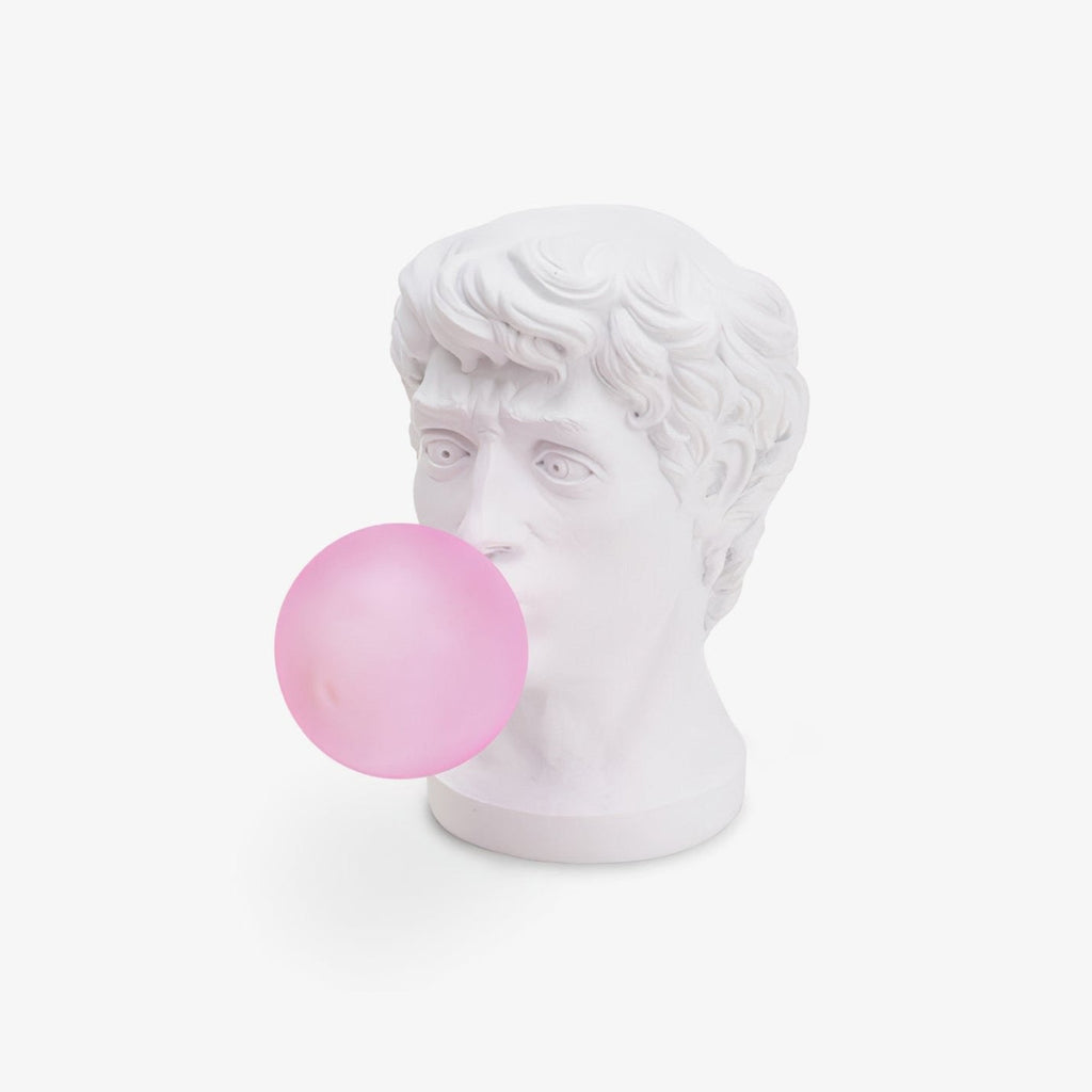 A Clérachic table lamp shaped as a white classical head blowing a pink glass bubble, on a plain white backdrop.