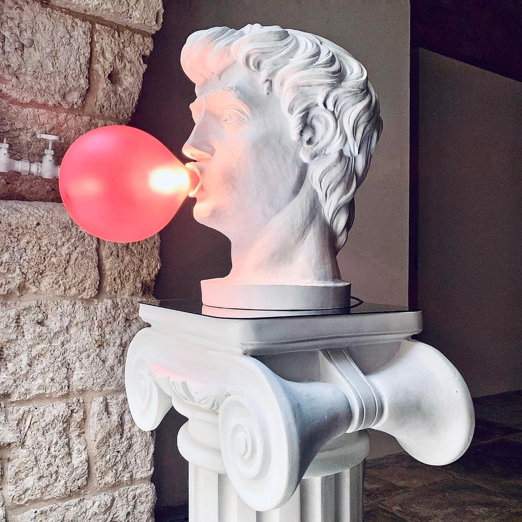 Seen in profile, a Clérachic bust lamp blows a glowing pink bubble, standing on a white Ionic column against a stone wall.