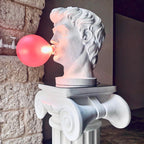 Seen in profile, a Clérachic bust lamp blows a glowing pink bubble, standing on a white Ionic column against a stone wall.