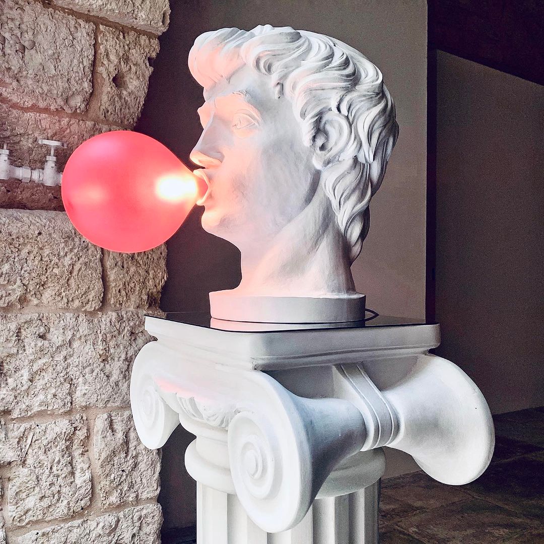 Seen in profile, a Clérachic bust lamp blows a glowing pink bubble, standing on a white Ionic column against a stone wall.