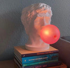 Lit warm, the bubble of this Clérachic bust lamp glows orange-pink as it stands on a stack of art books against a grey wall.