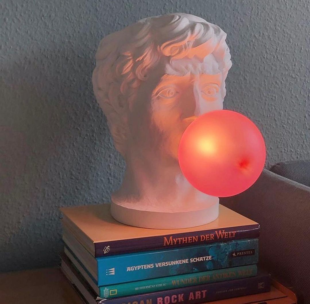 Lit warm, the bubble of this Clérachic bust lamp glows orange-pink as it stands on a stack of art books against a grey wall.