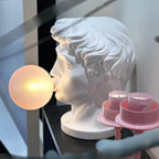 In profile on a dark shelf, a Clérachic white bust lamp blows a softly lit bubble, beside pink tea light holders.