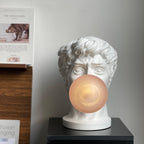 A white bust lamp from Clérachic with its peach-lit bubble over the mouth, on a black plinth against a pale wall.