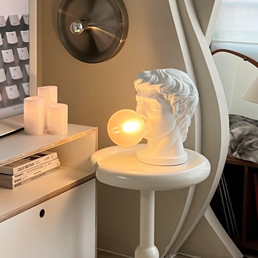 On a small round white table by a shelf of books, this Clérachic bust lamp glows with a warm amber bubble beside pillar candles.