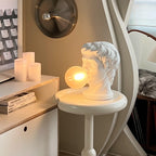On a small round white table by a shelf of books, this Clérachic bust lamp glows with a warm amber bubble beside pillar candles.