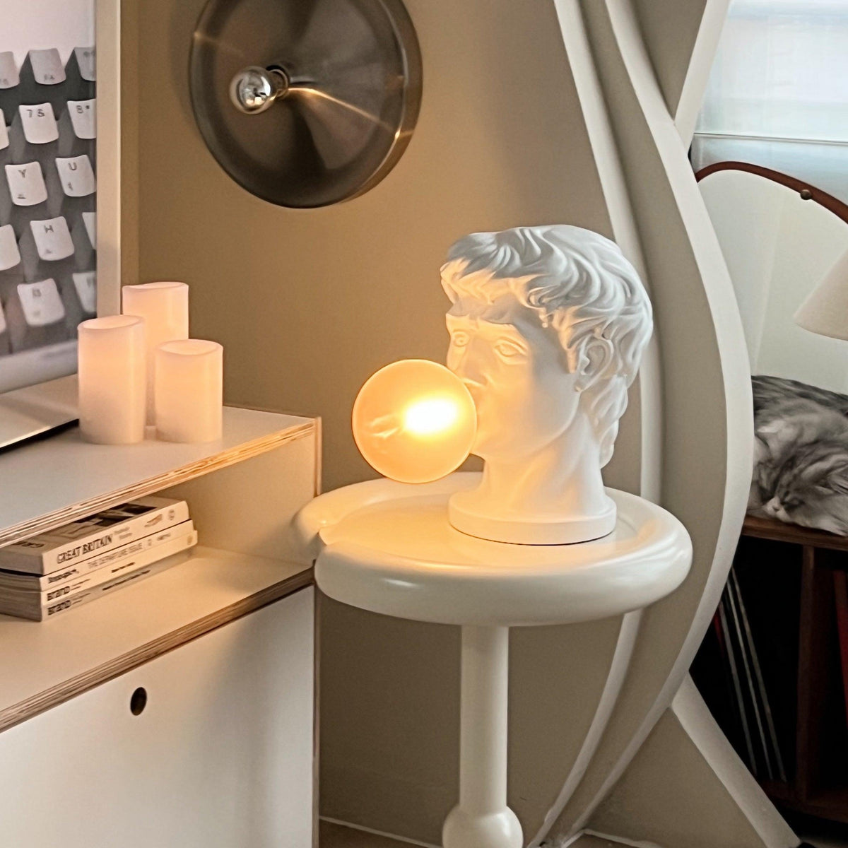 On a small round white table by a shelf of books, this Clérachic bust lamp glows with a warm amber bubble beside pillar candles.