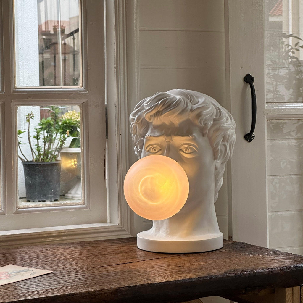 A Clérachic bust lamp stands on a dark wooden sill by a window, its lit bubble glowing a soft peach.