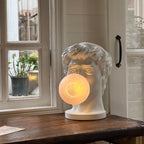 A Clérachic bust lamp stands on a dark wooden sill by a window, its lit bubble glowing a soft peach.