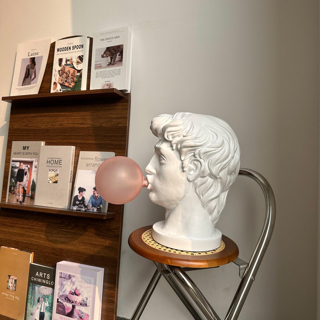 In profile on a wooden stool by a bookshelf, this Clérachic white bust lamp blows a soft pink bubble.