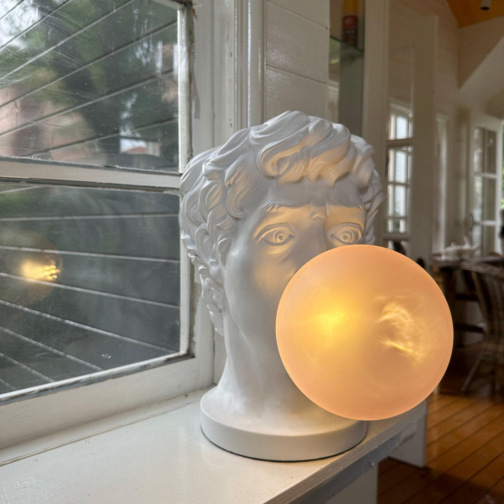 On a white windowsill, this Clérachic bust lamp has a large frosted bubble lit warm, with window blinds behind.