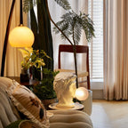 A small Clérachic bust lamp glows beside a chair and plants in a bright living room, its bubble lit a warm cream.