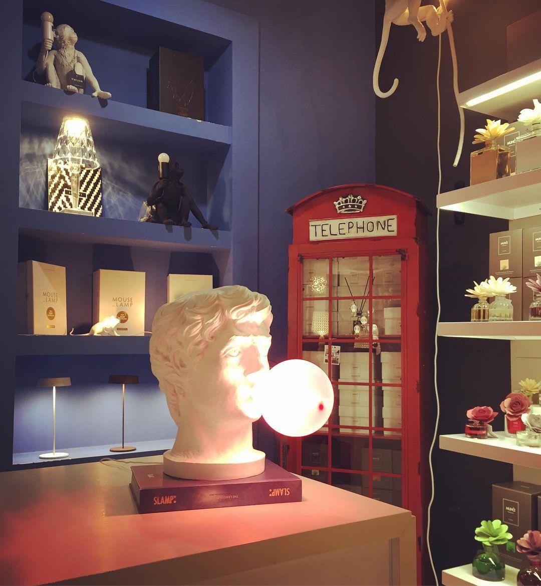 A Clérachic bust lamp on a stack of books in a blue-walled shop, its bubble lit warm beside a red telephone box.