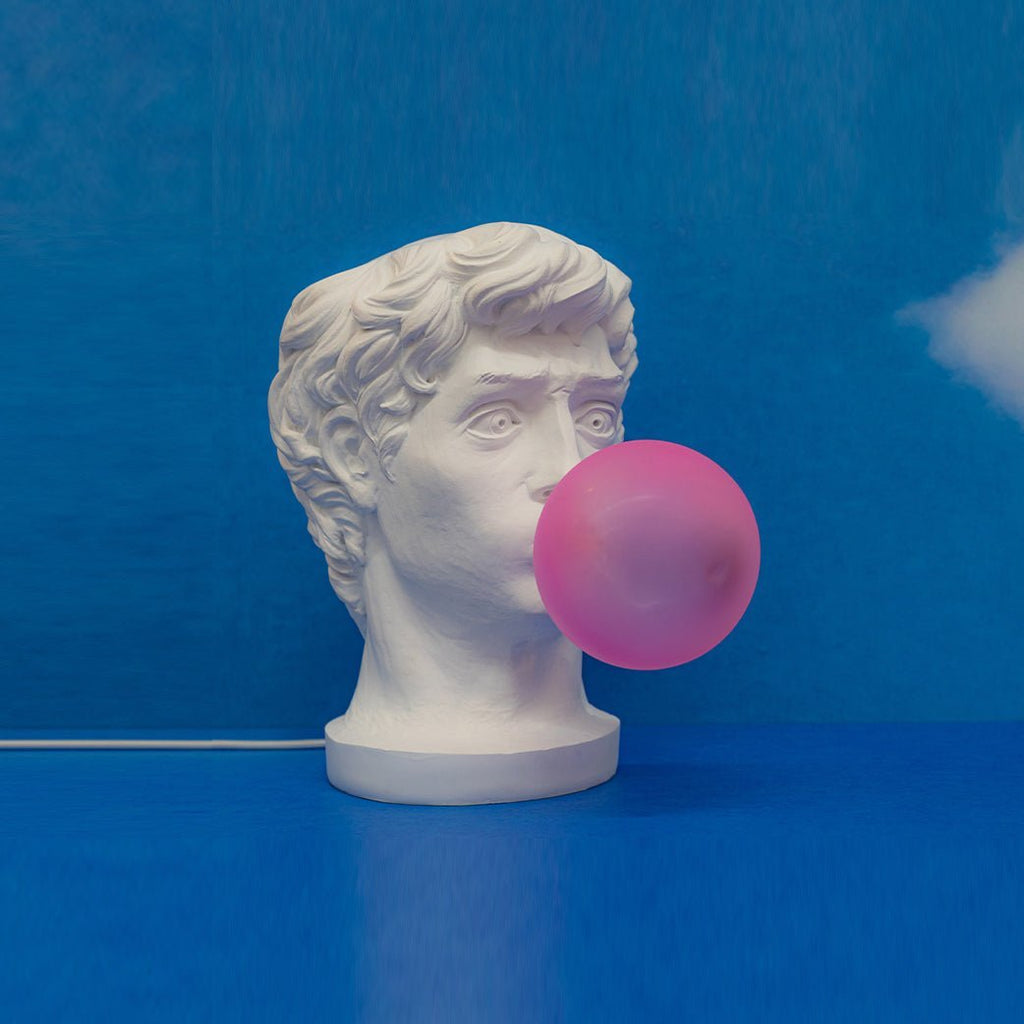 A Clérachic bust lamp with a pink bubble on a vivid blue backdrop and floor, its white cord trailing along the ground.