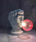 A Clérachic bust lamp with a glowing pink bubble stands on a marble surface in a dim room, with a soft pink shadow beside it.