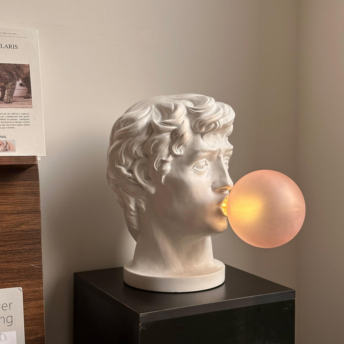 In profile on a black plinth, this Clérachic bust lamp has a lit peach bubble, with a framed notice on the wall behind.