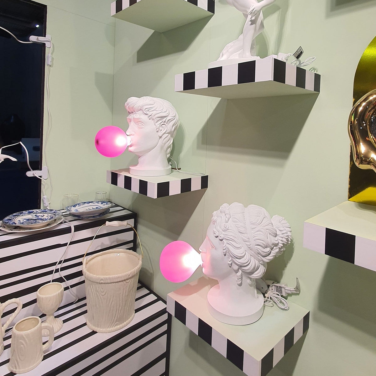 Two white bust lamps from Clérachic, one male and one female, glow with pink bubbles on black-and-white striped shelves against a pale green wall.
