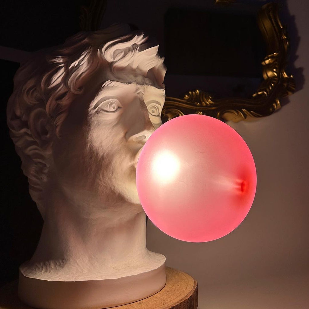 Close view of a Clérachic bust lamp with a bright pink-red bubble lit from inside, set against a dark gold-framed mirror.