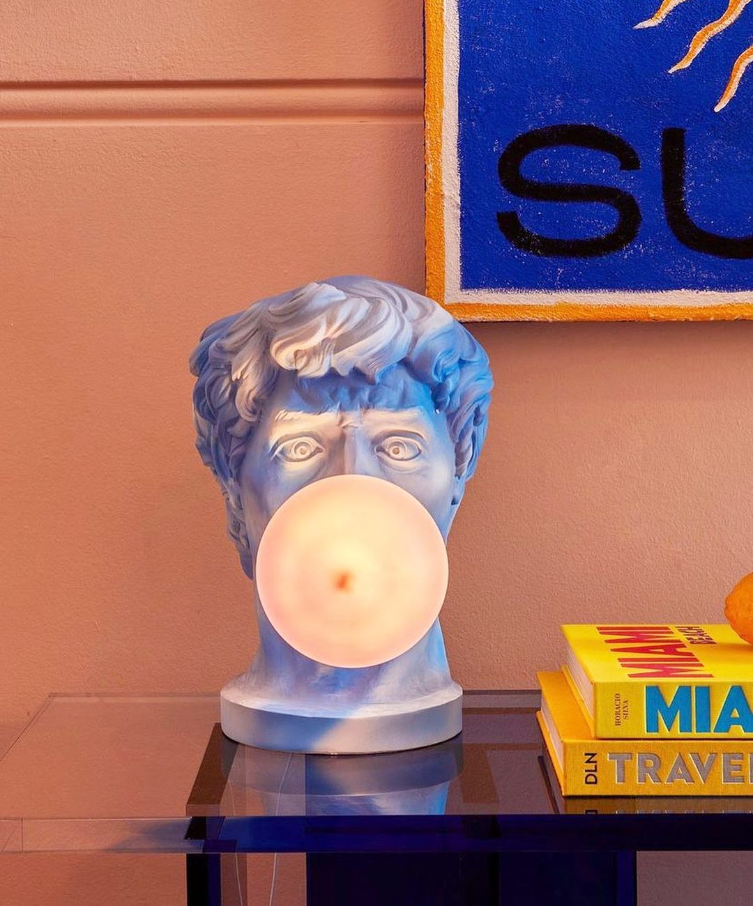 A Clérachic bust lamp stands on a glass table against a pink wall, its lit bubble cream and its curls tinted blue.