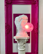 A Clérachic bust lamp with a glowing pink bubble stands on a white column inside a magenta ornate frame.