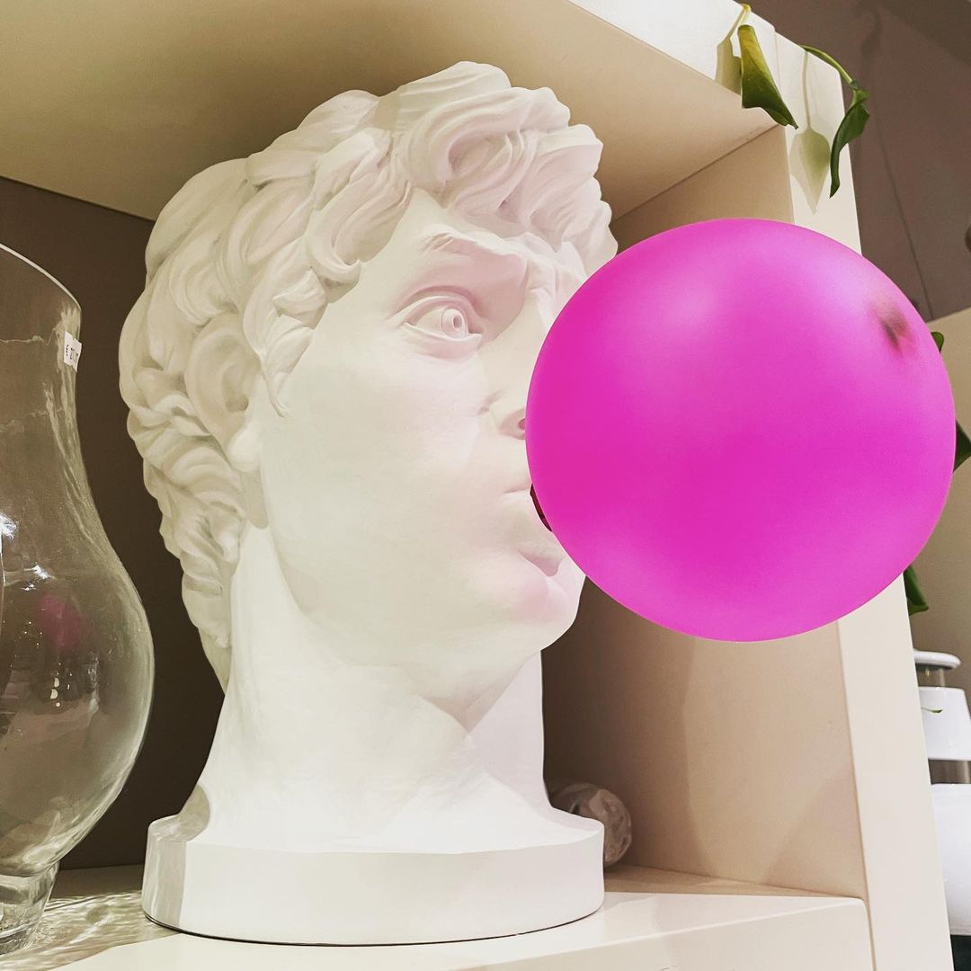 On a white shelf beside a tall glass vase, this Clérachic bust lamp has a bold magenta bubble over its mouth.