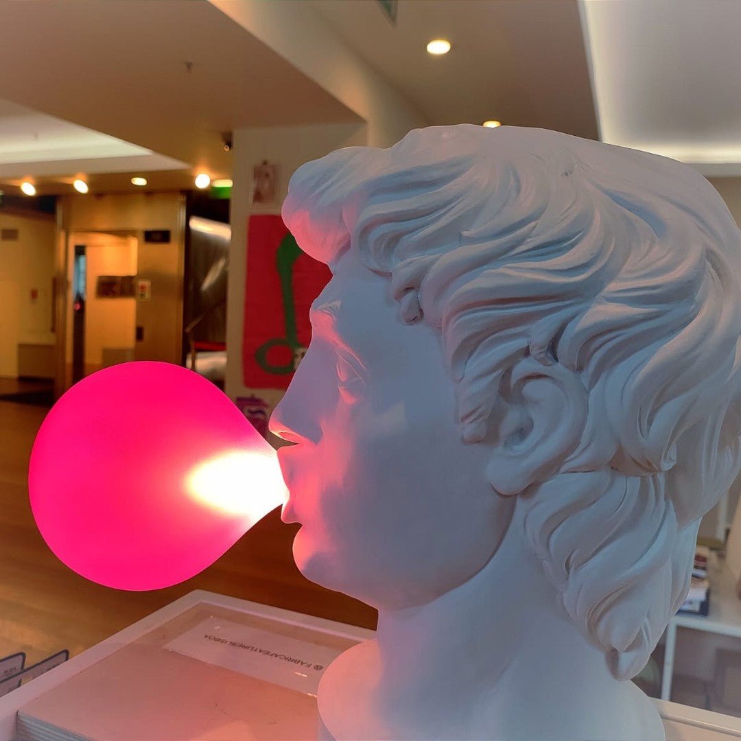 Close side view of a Clérachic bust lamp, its pink bubble glowing hot as the light shines out from the mouth, in a shop interior.
