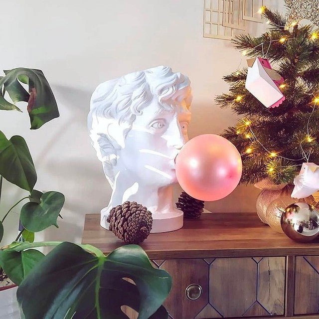 A Clérachic bust lamp on a wooden console, its pink bubble unlit, beside a monstera and a Christmas tree with warm lights.