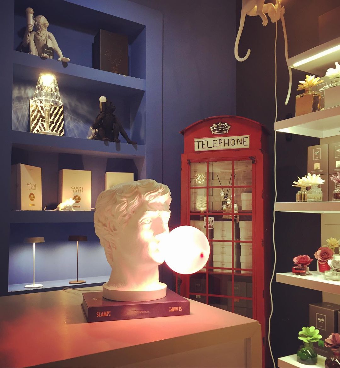 A Clérachic bust lamp with a lit bubble sits on a stack of books in a blue shelved room, with a red telephone box beyond.