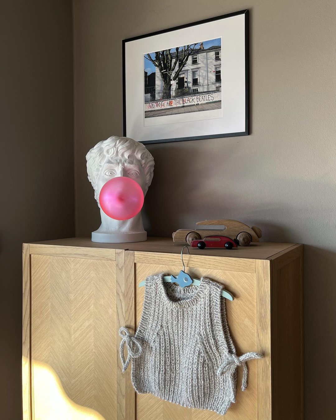 A Clérachic bust lamp on a pale wooden cabinet, its pink bubble unlit, with a small toy car and a knitted vest hanging beside it.