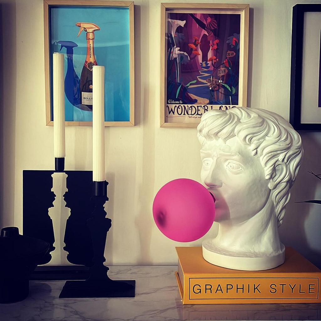 A Clérachic bust lamp on a black-and-white shelf with a gold book, its pink bubble unlit, beside two tall candles and framed prints.