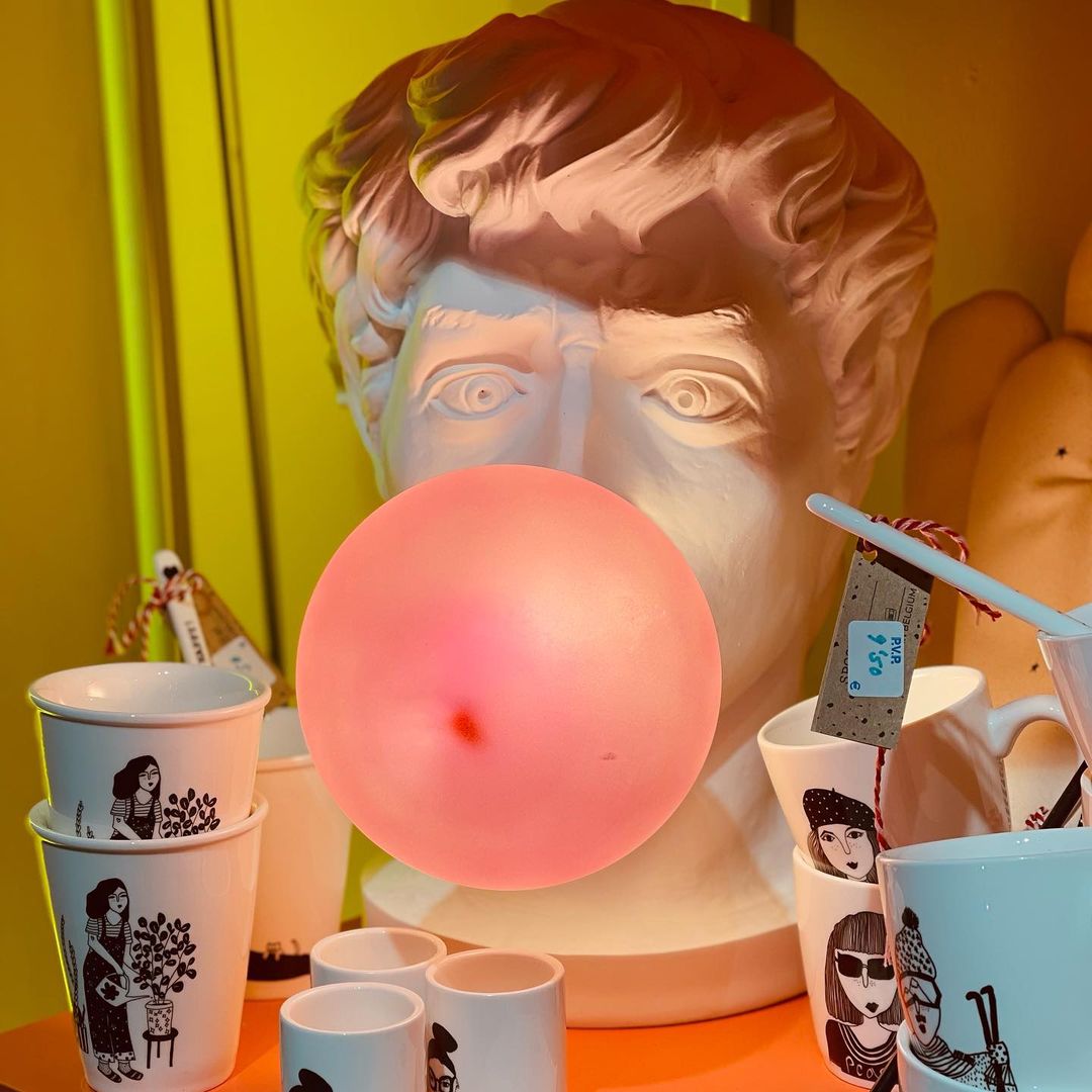 A Clérachic bust lamp and its glowing coral bubble fill the frame, with stacked mugs and a bright yellow wall around it.