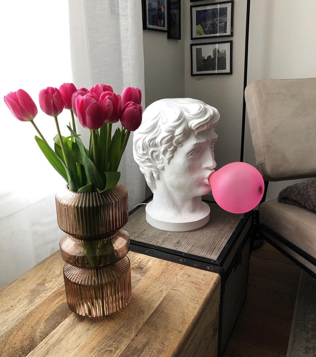 A Clérachic bust lamp stands on a wooden table by a vase of pink tulips and a grey armchair, its bubble unlit.
