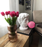A Clérachic bust lamp stands on a wooden table by a vase of pink tulips and a grey armchair, its bubble unlit.