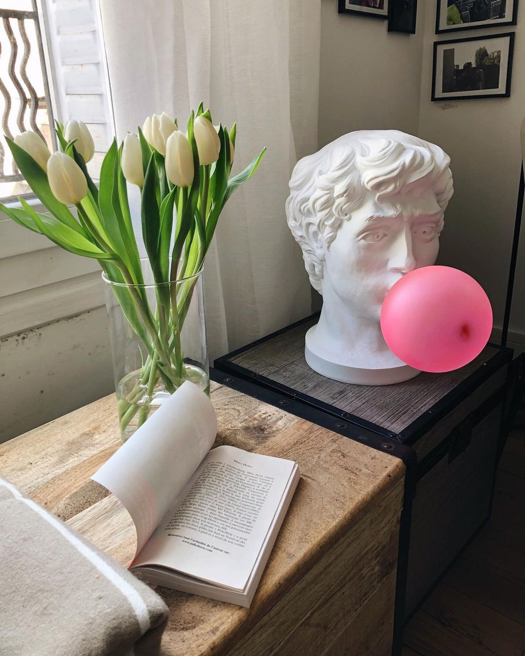 Beside a vase of white tulips and an open book, this Clérachic bust lamp has an unlit pink bubble on a rustic wooden table.