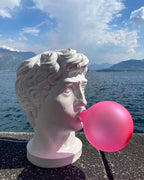 Outdoors on a stone ledge by a lake and mountains, this Clérachic bust lamp blows a bright pink bubble under a blue sky.